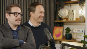 Photo of Dr George Ward and Prof Jan-Emmanuel De Neve in the Working on Wellbeing podcast studio. Both are looking off to the right, and on the shelf behind them is a copy of the 2024 World Happiness Report, which has an orange cover.