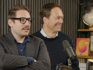 Photo of Dr George Ward and Prof Jan-Emmanuel De Neve in the Working on Wellbeing podcast studio. Both are looking off to the right, and on the shelf behind them is a copy of the 2024 World Happiness Report, which has an orange cover.