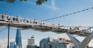 A photo of the Millennium Bridge in London. The Shard is pictured behind the bridge, and there are many pedestrians crossing the bridge. The sky is very clear and blue, and there are green leaves on the trees.
