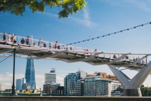 A photo of the Millennium Bridge in London. The Shard is pictured behind the bridge, and there are many pedestrians crossing the bridge. The sky is very clear and blue, and there are green leaves on the trees.